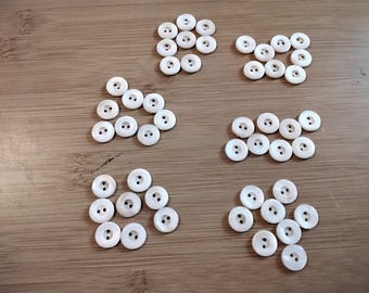 May include: A collection of white, round buttons with two holes, arranged in small groups on a wooden surface. The buttons are evenly spaced and appear to be made of a smooth, possibly pearlescent material. Ideal for sewing and craft projects.