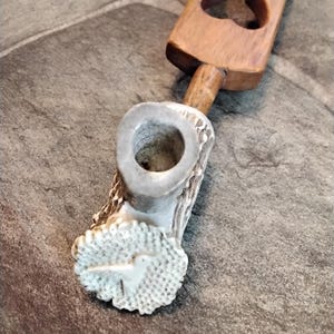 May include: A handcrafted pipe featuring a light brown wooden handle with heart-shaped cutouts and a white, textured bowl made from antler. The bowl has a carved bird design. The pipe is designed for smoking.