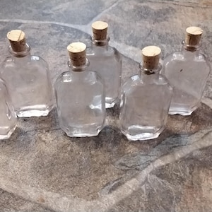 May include: Six clear glass bottles with cork stoppers. The bottles are all the same size and shape, and they are arranged in a row on a light brown surface.