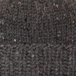 May include: Close-up of a dark grey knitted hat with a ribbed brim. The hat has a textured appearance with flecks of white and other colours throughout the yarn. The knit pattern is visible, showcasing the craftsmanship of the hat.