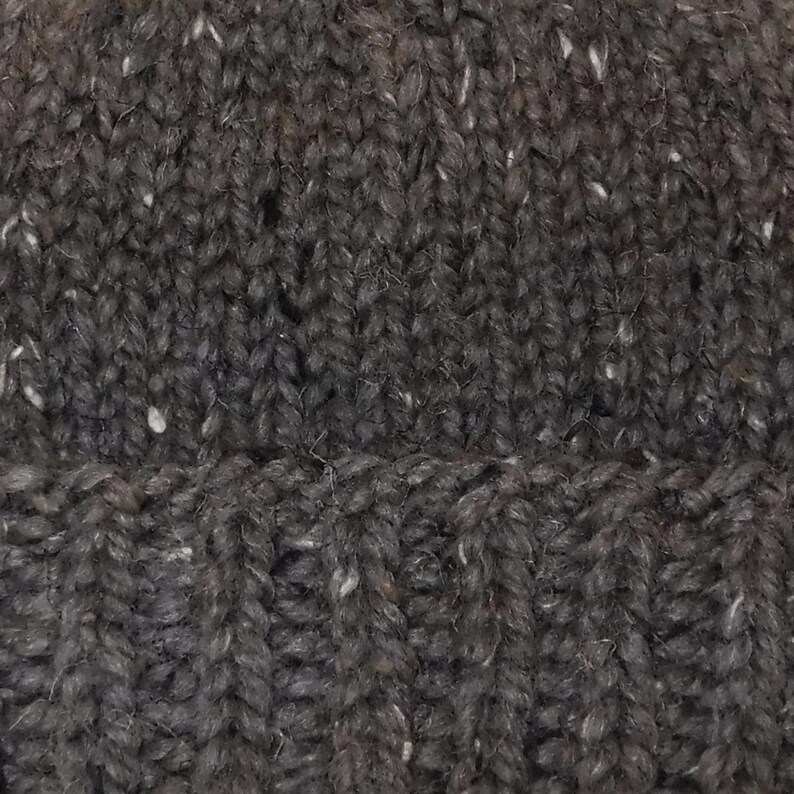 May include: Close-up of a dark grey knitted hat with a ribbed brim. The hat has a textured appearance with flecks of white and other colours throughout the yarn. The knit pattern is visible, showcasing the craftsmanship of the hat.