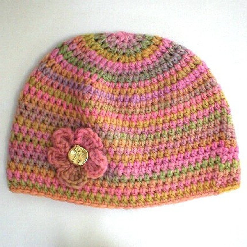 May include: A crocheted hat in shades of pink, green, and brown with a flower embellishment. The flower has a gold button in the center.