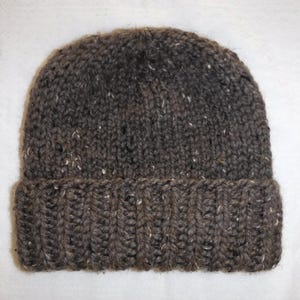 May include: A dark brown knitted beanie hat with a folded-up brim. The yarn has a textured appearance with white and tan flecks. The ribbed brim offers a close fit. Ideal for keeping warm in cold weather.