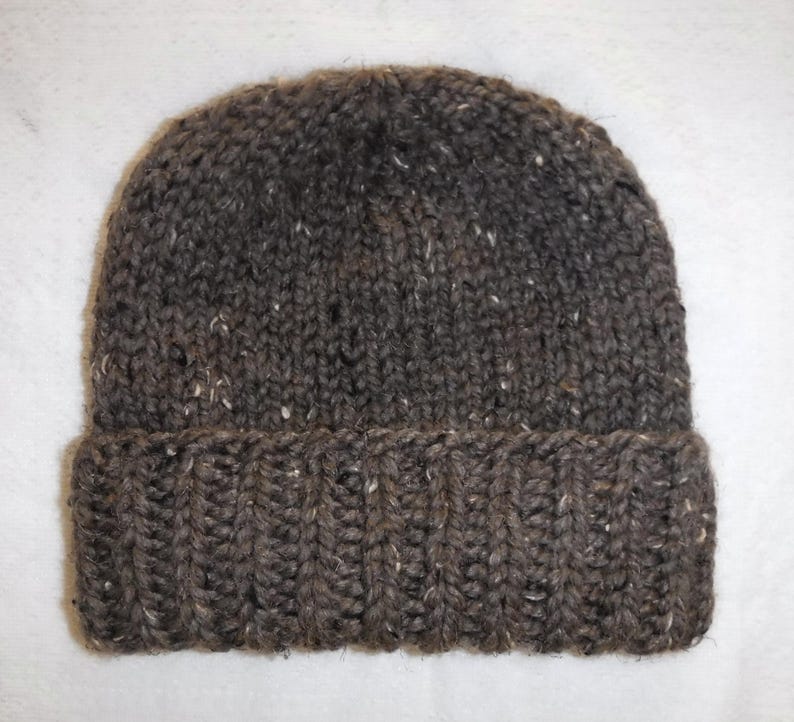 May include: A dark brown knitted beanie hat with a folded-up brim. The yarn has a textured appearance with white and tan flecks. The ribbed brim offers a close fit. Ideal for keeping warm in cold weather.