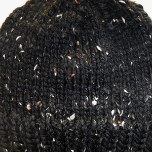 Puede incluir: Primer plano de un gorro de punto gris oscuro con motas blancas y marrones. El gorro est&aacute; hecho de un hilo grueso y texturizado, y el patr&oacute;n de punto es claramente visible. El gorro es un accesorio de invierno.