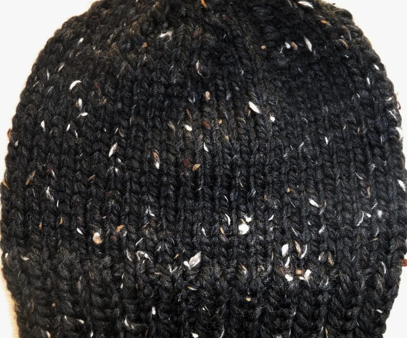 Puede incluir: Primer plano de un gorro de punto gris oscuro con motas blancas y marrones. El gorro est&aacute; hecho de un hilo grueso y texturizado, y el patr&oacute;n de punto es claramente visible. El gorro es un accesorio de invierno.