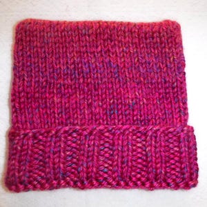 Op de afbeelding: Een handgebreide beanie-muts in tinten fuchsia, roze en paars. De muts heeft een geribbelde rand en een getextureerde gebreide body. Het garen heeft vlekjes blauw en geel. Deze warme muts is perfect voor koud weer.