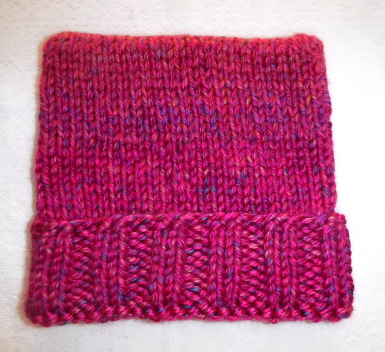 Op de afbeelding: Een handgebreide beanie-muts in tinten fuchsia, roze en paars. De muts heeft een geribbelde rand en een getextureerde gebreide body. Het garen heeft vlekjes blauw en geel. Deze warme muts is perfect voor koud weer.