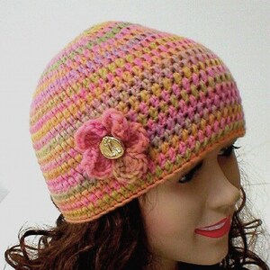 May include: A crocheted hat with a pink flower and a gold button. The hat is made with a multi-colored yarn in shades of pink, orange, and green.