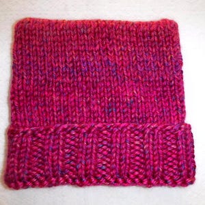 Op de afbeelding: Een handgebreide beanie in tinten fuchsia, magenta en paars. De hoed heeft een geribbelde rand en een getextureerde body van tricotsteek. Het garen heeft subtiele vlekjes blauw en geel.