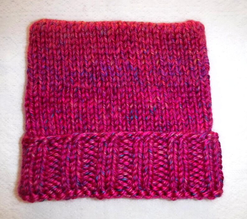 Op de afbeelding: Een handgebreide beanie in tinten fuchsia, magenta en paars. De hoed heeft een geribbelde rand en een getextureerde body van tricotsteek. Het garen heeft subtiele vlekjes blauw en geel.