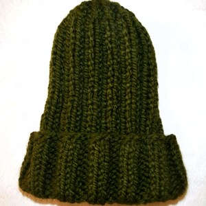 Op de afbeelding: Een donker olijfgroene gebreide beanie met een omgeslagen rand. De hoed is gemaakt van een dik, getextureerd garen en heeft een geribbeld patroon. De hoed wordt getoond tegen een witte achtergrond.