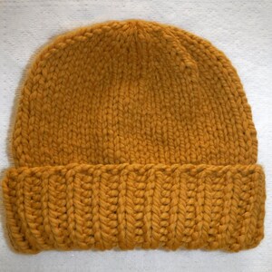 May include: Mustard yellow knit beanie hat. The hat is made of thick yarn and features a ribbed brim. The top portion of the hat has a simple knit pattern. This warm hat is perfect for cold weather.