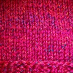Op de afbeelding: Close-up van een handgebreid item in roze en rode tinten. Het garen heeft spikkels van blauw en geel. De textuur van het breisel is zichtbaar, met een geribbeld patroon aan de onderkant.