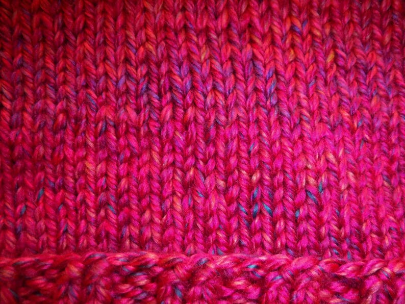 Op de afbeelding: Close-up van een handgebreid item in roze en rode tinten. Het garen heeft spikkels van blauw en geel. De textuur van het breisel is zichtbaar, met een geribbeld patroon aan de onderkant.