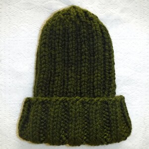 Op de afbeelding: Een donker olijfgroene gebreide muts met een omgeslagen rand. De muts is gemaakt van dik garen en heeft een geribbelde textuur. Een winteraccessoire.