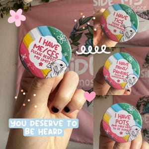 May include: A collection of four circular pin-back buttons featuring a colorful diagonal striped background. Each button displays a white cloud-shaped center with text and a line-art illustration of a rabbit. The text includes phrases like I HAVE ME/CFS, I HAVE TICS, I HAVE PANS/PANDAS, and I HAVE POTS, accompanied by requests for understanding or respect for limits.