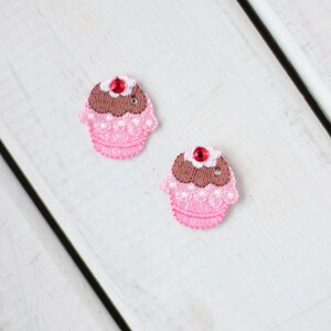 May include: Two pink cupcake-shaped fabric patches with brown icing and a red jewel-like cherry on top.