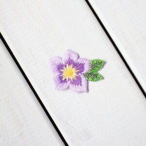 May include: An embroidered purple flower with a yellow centre and green leaves. The flower is on a white wooden background.