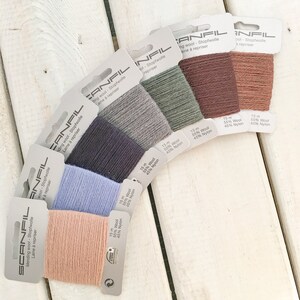 May include: A selection of SCANFIL mending wool spools in shades of pink, blue, grey, and brown. Each spool is labelled with the brand name and fibre content: 55% wool and 45% nylon. The spools are arranged in a semi-circle on a white wooden surface.