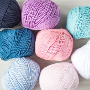 May include: Eight balls of yarn in various colours, including pink, blue, white, and purple. The yarn is arranged in a circle on a white surface.