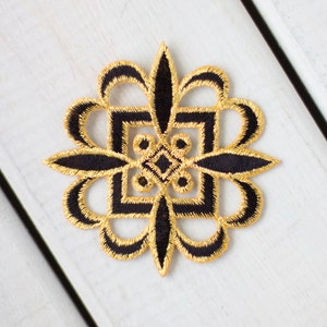 May include: A black and gold embroidered decorative patch with a geometric design. The patch is shaped like a flower with four petals.