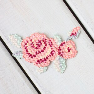 May include: A pink and white embroidered rose flower patch with green leaves. The patch is made with cross stitch embroidery.