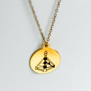 May include: Gold-colored pendant necklace with a circular charm. The charm features a geometric design with black lines and shapes. The necklace hangs against a white background.