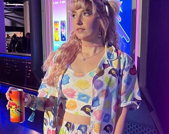 May include: A person wearing a colourful pajama set with a matching crop top and button-down shirt. The pajamas feature a pattern of various shapes and colours. The person is holding a can of a beverage.