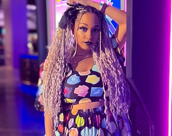 May include: A person in a matching crop top and trousers set with a vibrant, abstract pattern. The outfit has a black base with colourful shapes in blue, yellow, pink, and green. The setting is a room with neon purple lighting.