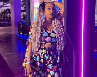 May include: A person wearing a matching crop top and wide-leg trousers set. The outfit features a black background with colourful, abstract shapes. The person is standing in front of a vending machine with a purple neon light.