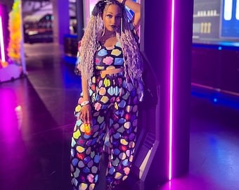 May include: A person in a matching crop top and trousers set with a vibrant, abstract pattern. The outfit has a black base with colourful shapes in blue, yellow, pink, and green. The setting is a room with neon purple lighting.