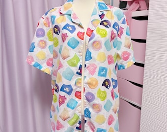 May include: A short-sleeved shirt on a mannequin. The white shirt is patterned with colourful, geometric shapes in shades of blue, yellow, pink, and purple. The shirt has a classic collar and is buttoned down the front.