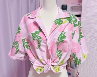 May include: A light pink shirt with a floral design featuring pink flowers and green leaves. The shirt is tied at the waist, creating a casual look. The short-sleeved shirt is displayed on a mannequin, showcasing a summery style.