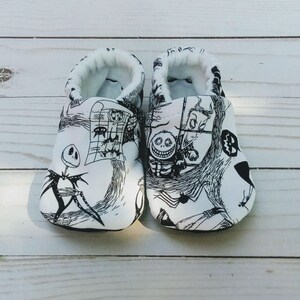 May include: A pair of white baby booties with a black and white print of characters from the Nightmare Before Christmas movie. The print includes Jack Skellington, Sally, and Oogie Boogie.