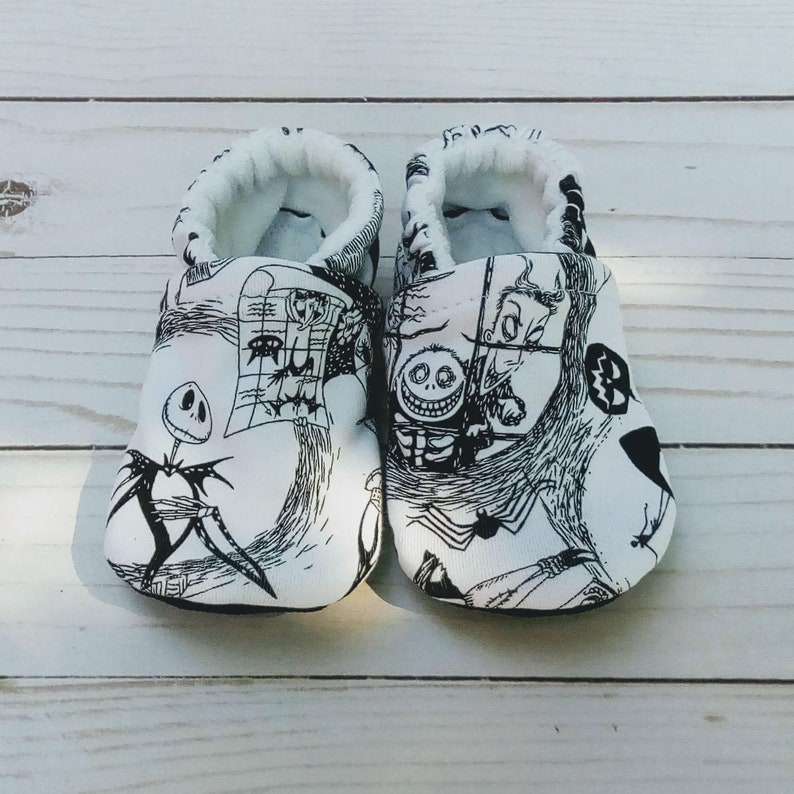 May include: A pair of white baby booties with a black and white print of characters from the Nightmare Before Christmas movie. The print includes Jack Skellington, Sally, and Oogie Boogie.