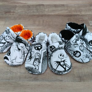 May include: Four pairs of black and white baby booties with a Nightmare Before Christmas theme. The booties have a white background with black illustrations of characters from the movie. Two pairs have orange trim and two pairs have black trim.