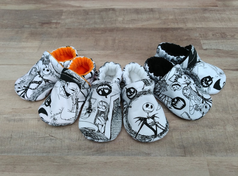 May include: Four pairs of black and white baby booties with a Nightmare Before Christmas theme. The booties have a white background with black illustrations of characters from the movie. Two pairs have orange trim and two pairs have black trim.