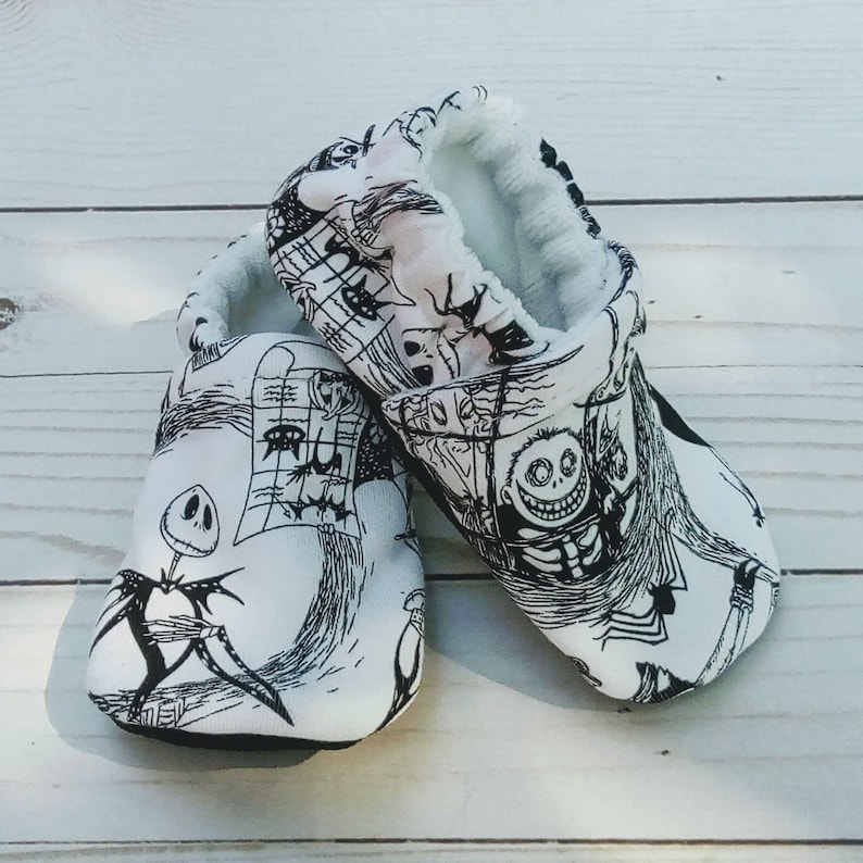 May include: A pair of white baby booties with a black and white print of characters from the Nightmare Before Christmas movie. The booties have a soft, fuzzy lining.