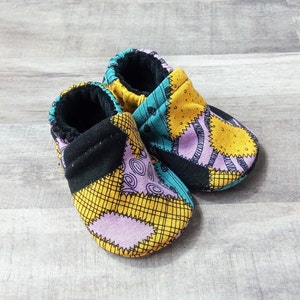 May include: A pair of baby booties with a patchwork design featuring black, yellow, purple, and blue fabric. The booties have a black cuff and a soft, fuzzy lining.