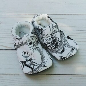 May include: A pair of white baby shoes with black and white illustrations of characters from the Nightmare Before Christmas movie. The shoes have a soft, comfortable fabric and are perfect for little feet.