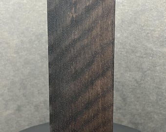 May include: A rectangular, dark brown wooden block with a visible wood grain pattern. The block is standing upright against a neutral grey background. The wood grain has a striped appearance.
