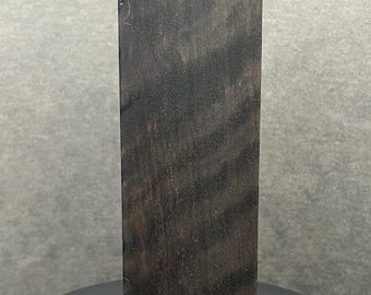 May include: A rectangular block of dark brown wood with a natural grain pattern. The wood has a smooth, polished surface and a slightly rounded top edge. The block is standing upright on a dark surface.