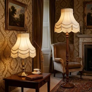 May include: Two antique lamps with cream-colored shades and fringe trim. One is a table lamp on a dark wood table, the other is a floor lamp. The room has patterned wallpaper, a fireplace, and a wingback chair, creating a vintage aesthetic.