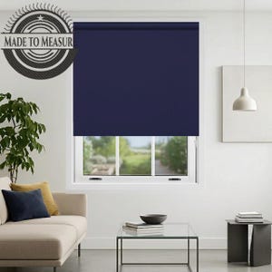 May include: A navy blue roller shade is installed over a window. The shade is pulled down, covering the window. The words "MADE TO MEASUR" are printed on a circular emblem in the upper left corner. The room has a sofa, coffee table, and side table.