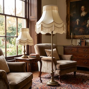 May include: Two lamps with cream-colored shades and fringe trim. One is a table lamp, the other a floor lamp. Both have ornate brass bases. The lamps are in a room with a large window, two armchairs, and a painting.