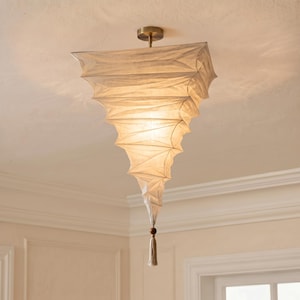 May include: A ceiling-mounted pendant light fitting featuring a cream-coloured, pleated fabric shade with a tapered, inverted pyramid shape. The light is suspended by a brass-toned metal mount and finished with a small wooden bead and fabric tassel.
