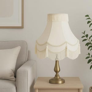 May include: Cream-colored table lamp with a fringed lampshade and a brass-colored base. The lampshade has a scalloped design with fringe and decorative trim. The lamp is on a light-colored wooden table.