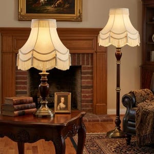 May include: Two elegant lamps with cream-colored shades and decorative fringe. One is a table lamp, the other a floor lamp. Both have dark wood bases and ornate brass accents. The lamps are in a room with a fireplace and antique furniture.