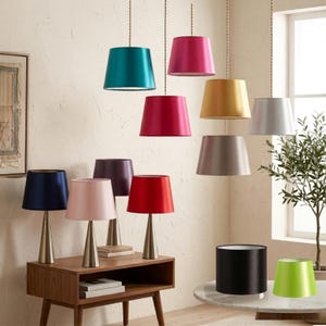 May include: A collection of colorful lampshades and table lamps. The lampshades are in various colors, including teal, pink, gold, silver, navy, purple, red, black, and green. The table lamps have metallic bases. The lampshades are made of a smooth, satin-like material.